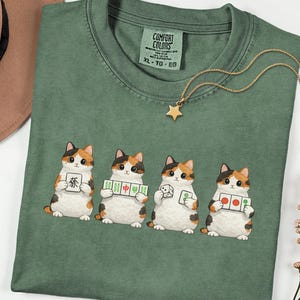 May include: Green t-shirt with a graphic of four calico cats holding Mahjong tiles. A gold star pendant necklace rests on the shirt. The shirt has a Comfort Colors tag and is size XL-TG-EG.