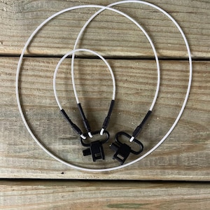 May include: Three white cords with black connectors and clasps are arranged in a circular pattern on a wooden surface. The cords are of varying sizes, with the largest forming a complete circle.