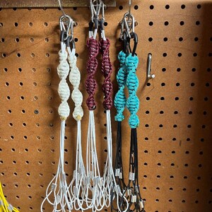 May include: Three macrame keychains in cream, burgundy, and turquoise hang from metal hooks. Each keychain features knotted beads and a loop at the bottom. The keychains are displayed against a pegboard background.
