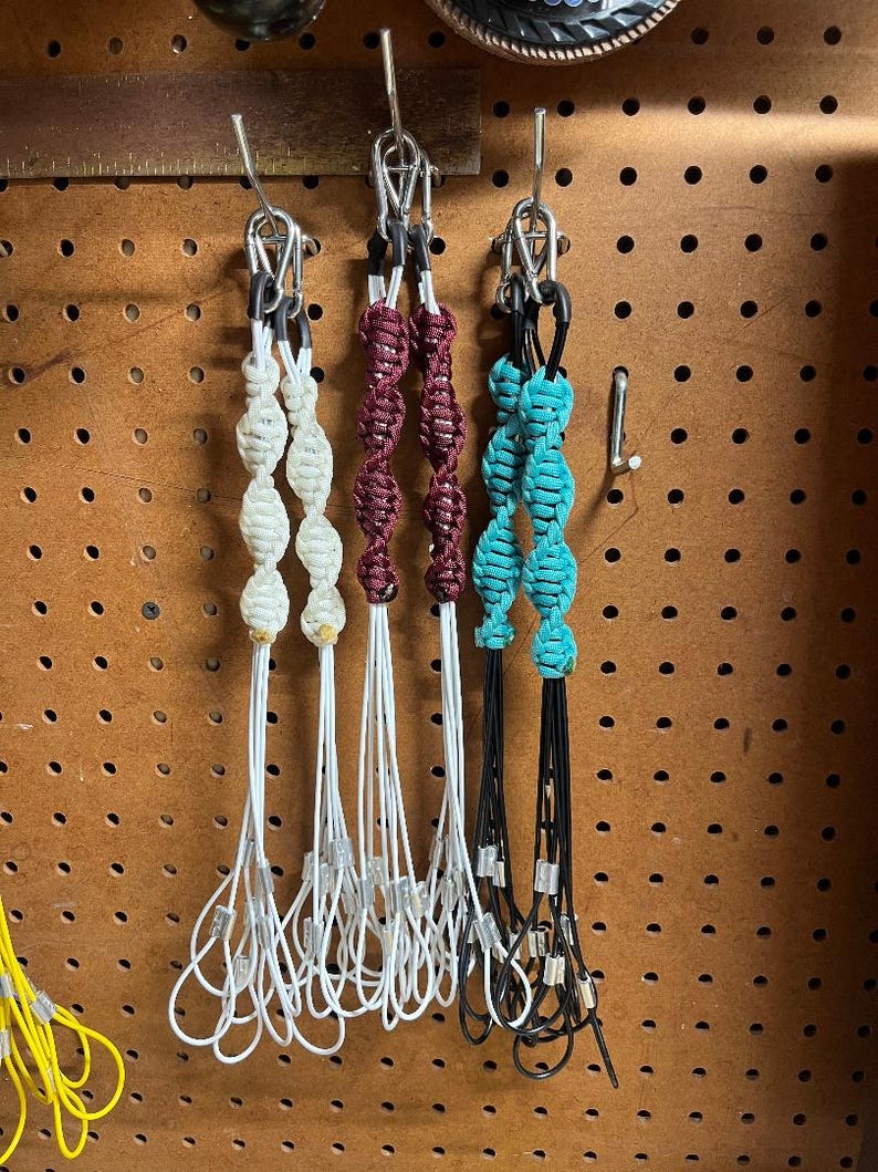 May include: Three macrame keychains in cream, burgundy, and turquoise hang from metal hooks. Each keychain features knotted beads and a loop at the bottom. The keychains are displayed against a pegboard background.