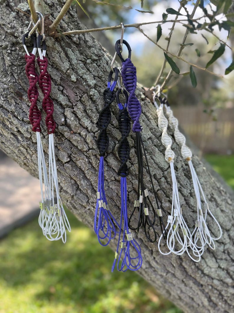 May include: A selection of macrame plant hangers in shades of burgundy, black, purple, and white. Each hanger is suspended from a metal hook and features a spiral design. The hangers are displayed on a tree branch, showcasing their intricate knotwork.