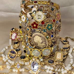 Puede incluir: Un frasco de vidrio transparente lleno de joyas doradas variadas, incluyendo collares, pulseras y anillos. La colección presenta camafeos, perlas de imitación, flores de esmalte de colores, pedrería y dijes en forma de luna de diversos tamaños.