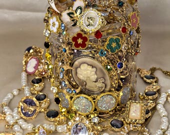 May include: A clear glass jar filled with assorted gold-tone jewelry including necklaces, bracelets, and rings. The collection features cameos, faux pearls, colorful enamel flowers, rhinestones, and moon-shaped charms in various sizes and designs.