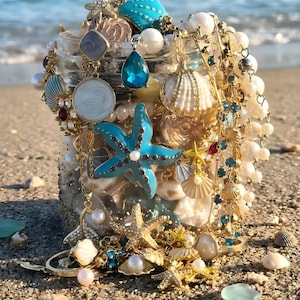 Puede incluir: Un tarro de cristal lleno de joyas y decoraciones con temática de playa. El tarro está lleno de conchas marinas, perlas y estrellas de mar turquesas. Cadenas y colgantes dorados con detalles azules y blancos caen en cascada, creando una estética costera.