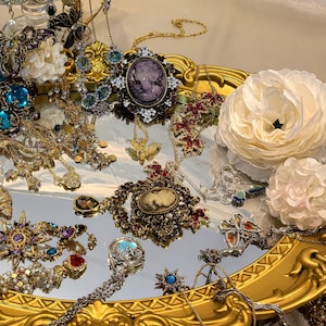 Op de afbeelding: Een assortiment vintage sieraden, waaronder kettingen, broches en oorbellen, gepresenteerd op een goudkleurige tray. De sieraden bevatten verschillende edelstenen, parels en ingewikkeld metaalwerk in goud- en zilverkleuren. Witte bloemen voegen een vleugje elegantie toe.