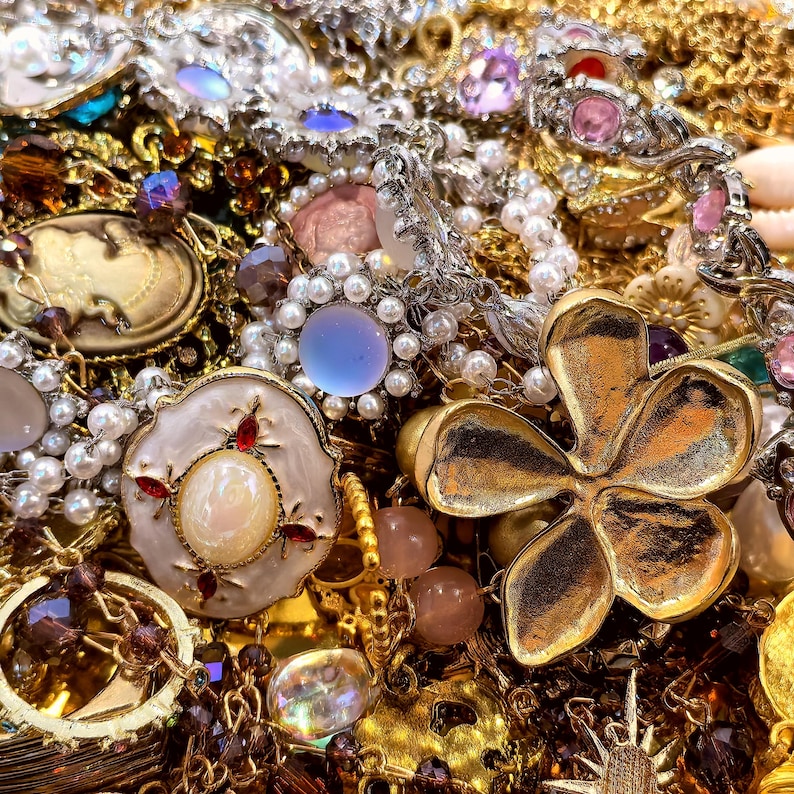 Op de afbeelding: Een close-up van een verzameling vintage sieraden, waaronder kettingen, armbanden en broches. De sieraden bevatten goud, zilver, parels en kleurrijke edelstenen. Een grote goudkleurige bloemvormige broche is prominent aanwezig.