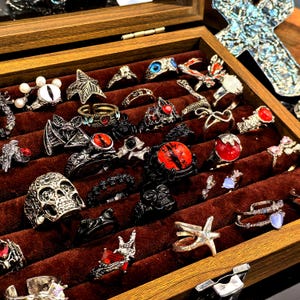 May include: A wooden display case filled with various rings. The rings feature diverse designs, including skulls, bats, and gemstones in red, blue, and white. The rings are made of silver and black metal.
