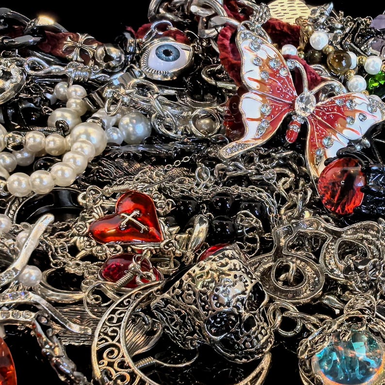 May include: A collection of various silver-toned necklaces, chains, and pendants. Notable pieces include a red heart with a cross, a butterfly with red and white wings, a skull, and a pearl necklace. The jewelry is arranged on a dark surface.