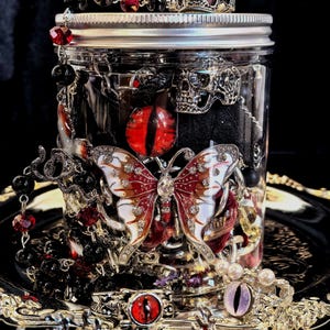 May include: A clear glass jar filled with gothic-themed jewelry and accessories. The jar is adorned with a silver skull ring, a butterfly pendant, and red and black beads. The contents include rings, beads, and decorative eye charms.