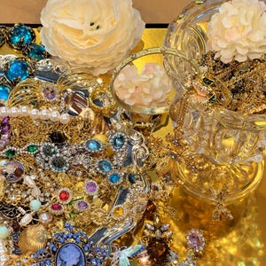 Op de afbeelding: Een verzameling gouden sieraden, waaronder kettingen, armbanden en oorbellen, met diverse edelstenen en parels. De sieraden zijn gerangschikt op een gouden oppervlak, met witte bloemen en een glazen kom.