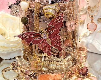 May include: A display of jewellery, including rings, necklaces, bracelets, and brooches, in gold, pink, and red hues. A large red butterfly brooch is central, surrounded by various decorative items. The jewellery is presented in a glass jar.