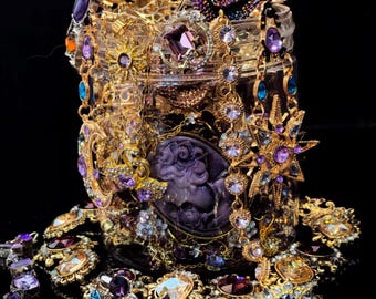 May include: A glass jar overflowing with ornate jewelry. Gold-toned necklaces, bracelets, and earrings are adorned with purple, blue, and clear gemstones. A purple rose and gold filigree accents the top, creating a luxurious display.