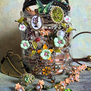 Pode incluir: Um frasco de vidro transparente transbordando com várias peças de joalheria. A joalheria inclui pulseiras, colares, anéis e brincos, com designs florais e de borboletas. A paleta de cores inclui detalhes dourados, verdes, rosa e brancos.