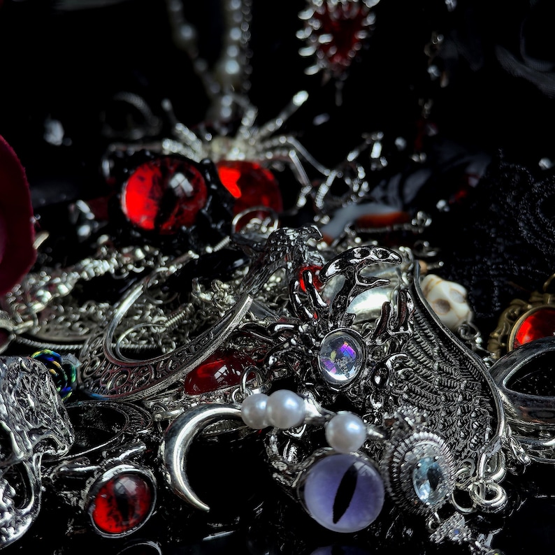 May include: An assortment of gothic-themed jewelry, including rings, pendants, and earrings, in silver and red. The collection features spider, eye, and crescent moon motifs, with pearl and gemstone accents. The items are arranged on a dark surface.