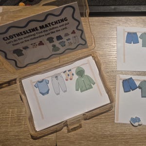 May include: A Clothesline Matching educational activity set featuring a clear plastic storage case with an instruction card titled CLOTHESLINE MATCHING, two printed cards showing clothing on a line, and several small cut-out clothing pieces.