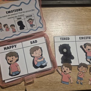 May include: An educational emotions matching game in a clear plastic case. The cards feature cartoon illustrations of a boy in a pink shirt and blue shorts expressing feelings like happy, sad, tired, and excited, with text labels and shadow silhouettes.