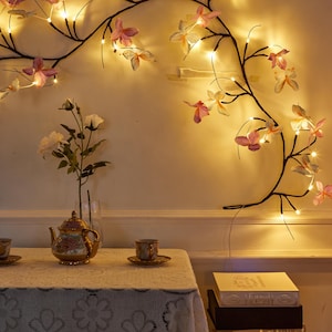 Puede incluir: Una rama decorativa montada en la pared con luces en forma de mariposa rosa y blanca. Una mesa está puesta con un mantel de encaje, una tetera, tazas y libros. La escena está iluminada por una luz cálida y suave.