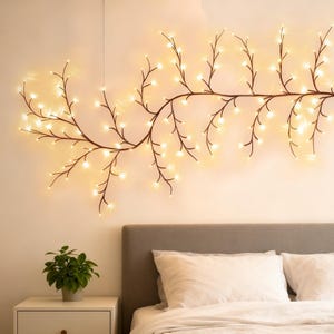 May include: A brown, branch-shaped wall light with small, warm white lights. The light is mounted on a light-coloured wall above a bed with white pillows and a bedside table with a potted plant.