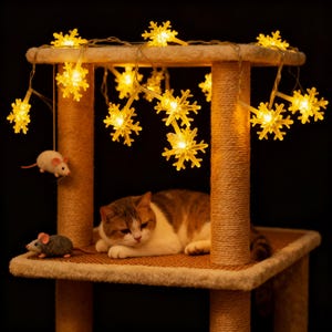 Op de afbeelding: Een kattenboom met een rustende kat, speelgoedmuizen en sneeuwvlokvormige lichtslingers. De kat is bruin, wit en oranje. De kattenboom heeft een beige platform met tapijt en met touw omwikkelde palen. De lichten zijn geel.