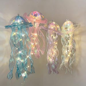 Pode incluir: Quatro decorações de medusas suspensas em tons de azul, rosa e branco iridescente. Cada medusa tem um topo transparente em forma de cúpula e tentáculos fluidos em forma de fita. Pequenas luzes são enfiadas dentro dos tentáculos.