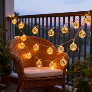 May include: A string of warm, glowing disco ball lights strung across a balcony railing. The lights are interspersed with a wicker chair with a white cushion. The background shows a dusky sky, suggesting an evening setting.