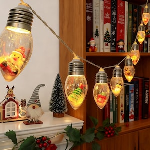 May include: A string of Christmas lights featuring clear, bulb-shaped ornaments. Each bulb houses a miniature festive scene, including Santa, a Christmas tree, and other holiday decorations. The lights are strung on a thin wire.