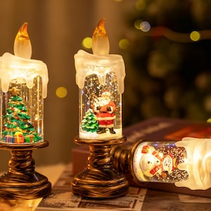 Könnte beinhalten: Weihnachtliche Kerzendekorationen mit warmem Schein. Die Kerzen haben ein klares, zylindrisches Design mit festlichen Szenen im Inneren, darunter ein Weihnachtsbaum und der Weihnachtsmann. Der Sockel ist dunkelbronzen.
