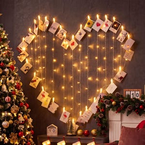 May include: A heart-shaped arrangement of warm white fairy lights with small photos clipped to the strands. The photos feature various images. A decorated Christmas tree and garland are visible in the background.