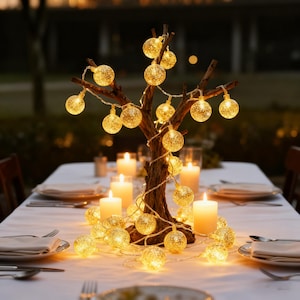 Op de afbeelding: Een tafeldecoratie met een houten boom versierd met bolvormige, gloeiende lichten. De warme, gouden lichten zijn zo gerangschikt dat ze op een boom lijken, waardoor een zachte gloed ontstaat. Verschillende witte kaarsen en couverts zijn op een wit tafelkleed geplaatst.