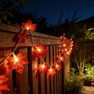 May include: A string of orange maple leaf lights draped over a wooden fence. Each leaf contains a small, warm white light, creating a cosy atmosphere. The background shows a dark night sky and some foliage.