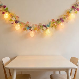 Puede incluir: Una guirnalda festiva de Pascua con luces en forma de huevo y coloridos acentos florales cuelga sobre una mesa y sillas blancas. La guirnalda presenta colores pastel y añade un toque primaveral a la habitación. Las luces brillan.
