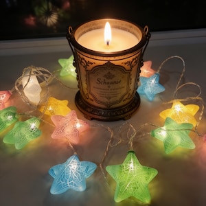 Peut inclure: Une bougie allumée dans un bougeoir décoratif avec le texte "Schaanie" et une guirlande lumineuse en forme d'étoiles colorées. Le bougeoir est de couleur bronze avec une poignée. Les étoiles sont roses, bleues, vertes et jaunes.