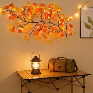 May include: A wall decoration of autumn leaves with warm white lights. A vintage lantern and a canvas duffel bag sit on a folding table. A framed print with text and botanical art hangs on the wall.