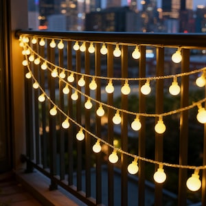 May include: String lights with warm white globe bulbs are strung across a black metal railing. The lights are illuminated, creating a cozy ambiance against a blurred cityscape background. The lights are perfect for decorating a balcony or patio.