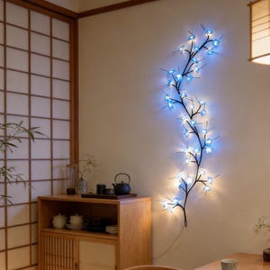 Può includere: Un'opera d'arte da parete in metallo nero a forma di ramo fiorito, adornata con luci a LED blu. Le luci sono disposte per imitare i fiori, creando un accento decorativo e illuminato. L'arte è montata su una parete di colore chiaro.
