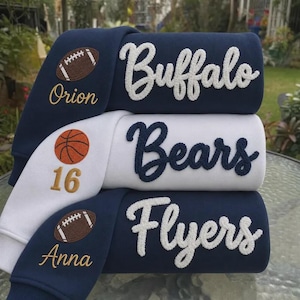 May include: Three stacked sweatshirts featuring chenille lettering. The top navy sweatshirt says Buffalo with a football, the middle white sweatshirt says Bears with a basketball and number 16, and the bottom navy sweatshirt says Flyers with a football.