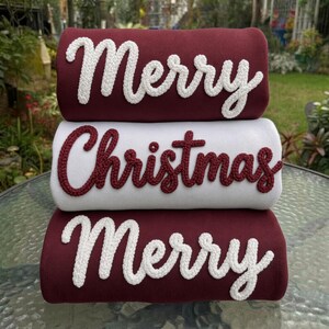 May include: A stack of three folded sweatshirts. The top and bottom sweatshirts are dark red with white embroidered text reading Merry. The middle sweatshirt is white with dark red embroidered text reading Christmas in cursive script.