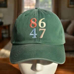 May include: A green baseball cap with the embroidered numbers 86, 6, 4, and 7 in various colors. The hat has a curved brim and a fabric strap closure. The hat is made of a soft, durable material.
