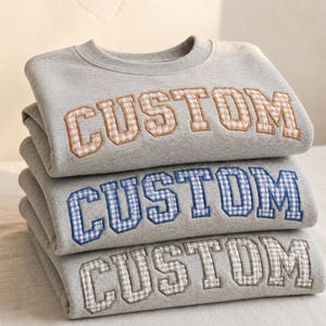 May include: A stack of three light gray crewneck sweatshirts, each featuring the word CUSTOM embroidered in a collegiate font. The lettering on each sweatshirt is filled with a different gingham check pattern in tan, blue, and gray colors.