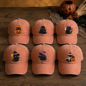 May include: Six orange baseball caps with embroidered designs. The top row features a ghost in a pumpkin, a ghost with a black cat, and a ghost holding a candle with flowers. The bottom row shows a ghost with a broom, a ghost on a skateboard, and a mummy ghost with a pumpkin bucket.