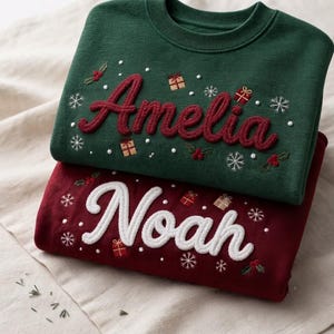 May include: A dark green sweatshirt with the name Amelia embroidered in red, and a dark red sweatshirt with the name Noah embroidered in white. Both feature embroidered snowflakes, small gift boxes, and holly berries across the front fabric.