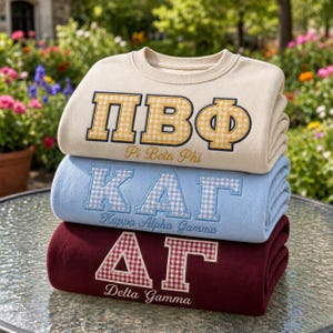 May include: A stack of three crewneck sweatshirts in cream, light blue, and maroon. Each features embroidered Greek letters with a gingham pattern fill and the corresponding organization name stitched in cursive script underneath the letters.