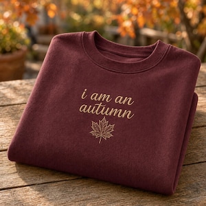 May include: A deep maroon crewneck sweatshirt featuring the embroidered text i am an autumn in a light tan cursive font, positioned above a detailed, single-line embroidered illustration of a maple leaf in the same light tan color.