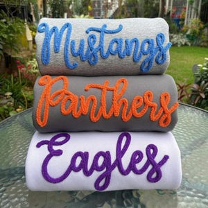 May include: Three folded sweatshirts stacked vertically. The top light gray sweatshirt features the blue text "Mustangs". The middle dark gray sweatshirt has orange text reading "Panthers". The bottom white sweatshirt displays purple text "Eagles".