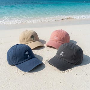 May include: Four baseball caps in navy blue, beige, dusty pink, and charcoal gray. Each cap features a small, simple white embroidered icon: a palm tree on the blue, a cactus on the beige, a rising sun on the pink, and a pine tree on the gray.