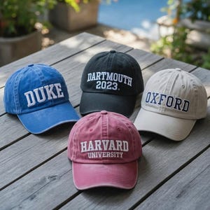May include: Four baseball caps in blue, black, pink, and tan are displayed on a wooden surface. Each cap features white embroidered text: "DUKE", "DARTMOUTH 2023", "OXFORD", and "HARVARD UNIVERSITY". The caps are arranged in a casual, outdoor setting.
