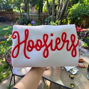 May include: A white rectangular pillow with the word "Hoosiers" embroidered in red cursive script. The pillow is held up in front of a blurred outdoor background with trees and foliage.