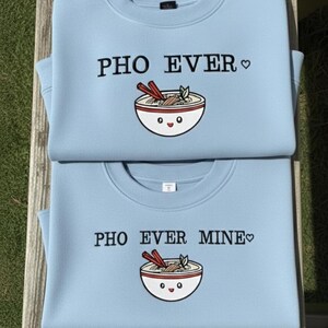 May include: Two light blue sweatshirts with the text "PHO EVER" and "PHO EVER MINE" in black. Each sweatshirt features a cartoon bowl of pho with chopsticks. The sweatshirts are folded and stacked on top of each other.