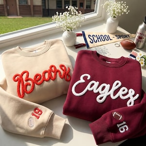 May include: A beige sweatshirt with red chenille lettering reading Bears and a maroon sweatshirt with white chenille lettering reading Eagles. Both feature embroidered footballs and numbers on the sleeves, with 19 on the beige and 16 on the maroon.