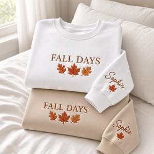 May include: A white sweatshirt and a beige sweatshirt, each featuring the embroidered text FALL DAYS above three maple leaves in shades of orange and brown. The sleeve of each sweatshirt is embroidered with the name Sophie and a single maple leaf.